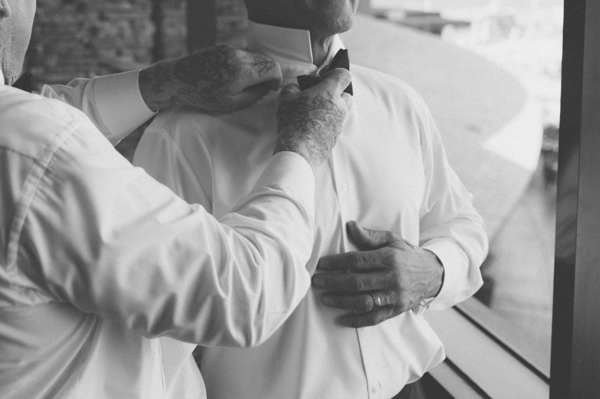 Groom getting ready — friend helping with the bow tie, candid moment captured by Manuel & Alma